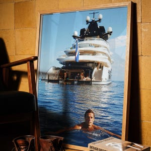 May include: A framed photograph depicting a large white yacht on blue water with a person swimming in the foreground. The image is displayed in a light wood frame, positioned near a pair of brown leather boots and a stack of books titled Design.