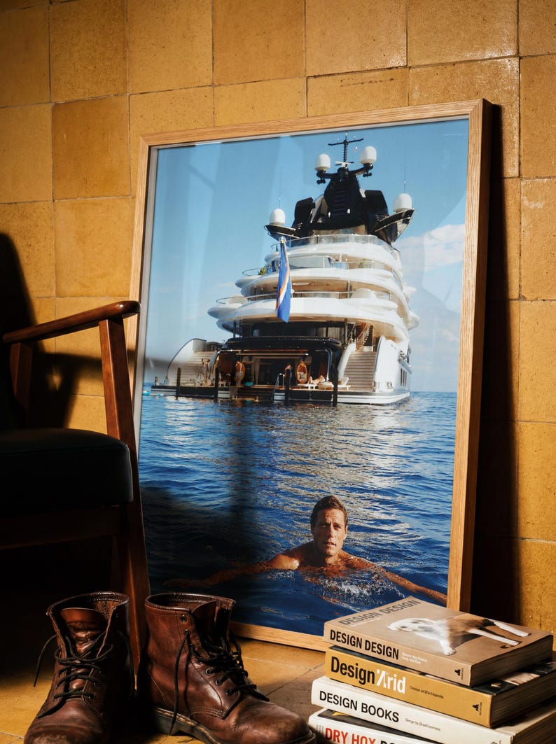 May include: A framed photograph depicting a large white yacht on blue water with a person swimming in the foreground. The image is displayed in a light wood frame, positioned near a pair of brown leather boots and a stack of books titled Design.