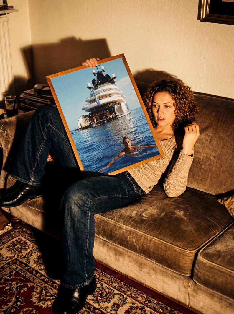 May include: A rectangular wooden-framed print featuring a large white yacht anchored in deep blue water with a person swimming in the foreground. The artwork captures a bright, sunny day at sea, showcasing the vessel's multi-level deck structure.