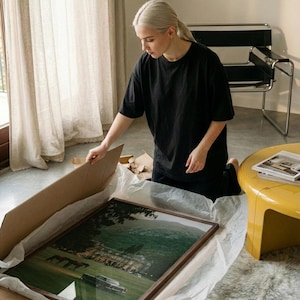 May include: A rectangular framed wall art print featuring a landscape scene with a large estate, green hills, and a vintage car parked on a lawn. The print is housed in a brown wooden frame and is partially wrapped in white tissue paper inside a cardboard box.