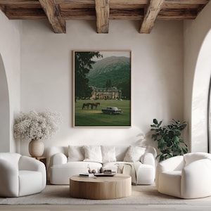 May include: A rectangular framed wall art print featuring a landscape scene with a large manor house, two horses, and a vintage car on a green lawn against a mountain backdrop. The artwork is displayed on a light-colored wall above a white sofa.