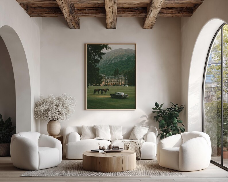May include: A rectangular framed wall art print featuring a landscape scene with a large manor house, two horses, and a vintage car on a green lawn against a mountain backdrop. The artwork is displayed on a light-colored wall above a white sofa.