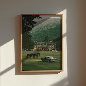 May include: A framed wall art print featuring a large estate building with a green lawn, two brown horses, and a black vintage car. The background shows a mountain range under a dark sky, all enclosed within a light-colored wooden frame.
