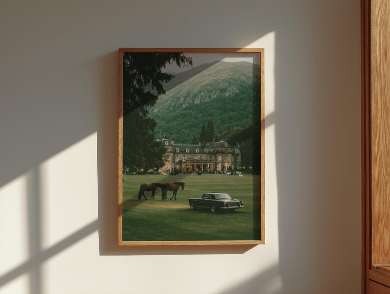 May include: A framed wall art print featuring a large estate building with a green lawn, two brown horses, and a black vintage car. The background shows a mountain range under a dark sky, all enclosed within a light-colored wooden frame.