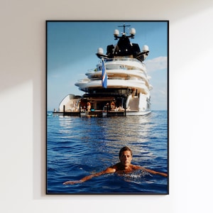 May include: A large white multi-deck yacht floats on deep blue ocean water. A person is swimming in the foreground, facing the camera. The yacht features a dark upper structure, multiple levels with railings, and a blue flag hanging from the stern.