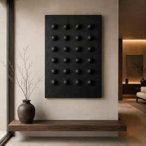 May include: A rectangular black wall art piece featuring a grid of twenty-four raised spherical protrusions. Beside it sits a dark brown ceramic vase containing thin, bare branches. Below the art is a long, horizontal wooden floating shelf.