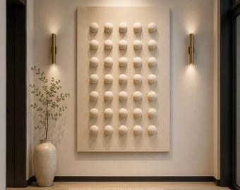 Op de afbeelding: Een groot rechthoekig wandkunstwerk met een raster van vijfendertig verhoogde crèmekleurige bollen. Het kunstwerk is bevestigd aan een neutrale muur tussen twee verticale goudkleurige wandlampen, met een hoge keramische vaas met een plant.
