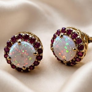 May include: A pair of round stud earrings featuring a central white opal with iridescent flashes, surrounded by a halo of small, deep red rubies set in yellow gold. The metal settings display a detailed, openwork lattice pattern on the sides.