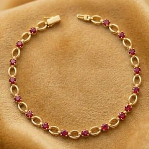 Könnte beinhalten: Ein goldenes Kettenarmband mit einer Abfolge von ovalen Gliedern und kleinen, runden, tiefroten Edelsteinen. Das Armband wird mit einem rechteckigen Kastenschloss verschlossen und bildet ein durchgehendes kreisförmiges Design.