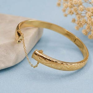May include: A gold-toned hinged bangle bracelet featuring an intricate engraved floral pattern across its surface. The bracelet includes a delicate attached safety chain that connects the two ends, securing the clasp mechanism firmly in place.