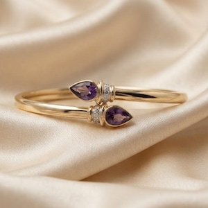 May include: A gold bypass bangle bracelet featuring two pear-shaped purple amethyst gemstones at the terminals. Each gemstone is accented by a small band of pavé-set clear stones, creating a refined look with a polished gold finish.