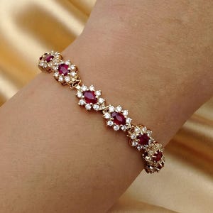 May include: A gold bracelet featuring a series of oval-cut deep red rubies, each surrounded by a halo of small, clear, round-cut white stones. The links are connected in a repeating floral-inspired pattern, creating a continuous band of gemstones.