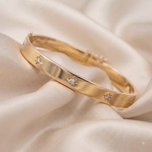 May include: A gold hinged bangle bracelet featuring a smooth, polished finish and multiple star-shaped settings. Each star motif contains a small, clear, round-cut gemstone at its center, evenly spaced along the outer surface of the metal band.