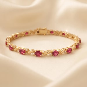 May include: A gold-tone bracelet featuring a series of oval-cut red gemstones alternating with small clear stones. The metal links have a twisted, serpentine design connecting each stone setting, and the piece includes a secure box clasp closure.