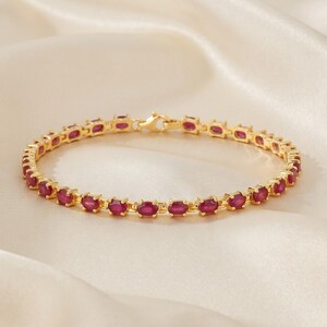 May include: A gold-toned bracelet featuring a continuous row of oval-shaped, deep red gemstones set in individual metal frames. The piece includes a secure lobster claw clasp mechanism, creating a circular design with uniform links throughout.