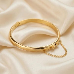 Puede incluir: Una pulsera rígida articulada de tono dorado pulido con una superficie lisa y redondeada. La pulsera cuenta con un mecanismo de cierre seguro y una delicada cadena de seguridad a juego, sujeta a ambos lados de la bisagra para evitar pérdidas.