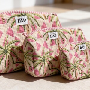 Peut inclure: Trois trousses matelassées à fermeture éclair de tailles décroissantes, avec un fond crème et un motif répétitif de triangles roses et de palmiers verts. Chaque trousse porte une étiquette rectangulaire blanche avec le texte DAP en noir.