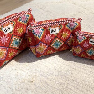 May include: Three red zippered pouches in varying sizes, each featuring a colourful geometric star and diamond pattern. The fabric has a quilted texture, and each pouch includes a white rectangular label with the black text DAP sewn on the front.