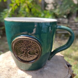 Könnte beinhalten: Ein glänzender, dunkelgrüner Keramikbecher mit einem kreisförmigen Medaillon auf der Vorderseite. Das Medaillon zeigt ein erhabenes, detailliertes Relief eines Baumes mit komplexen Ästen und Wurzeln in einem kontrastierenden Braunton.