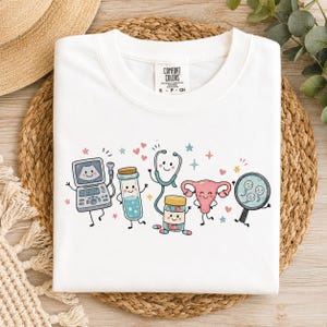 May include: A white t-shirt featuring a graphic print of cartoon medical items including an ultrasound machine, a test tube, a stethoscope, a pill bottle, a uterus, and a magnifying glass with sperm cells, all with smiling faces and hearts.