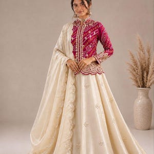 May include: A magenta long-sleeved jacket with a white bandhani pattern, a high collar, and scalloped hem, paired with a cream-colored floor-length skirt featuring scattered floral embroidery and a matching cream dupatta with a scalloped edge.