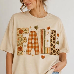 May include: A beige short-sleeve t-shirt featuring the word FALL printed in large block letters. Each letter has a unique pattern including floral, orange and white gingham, vertical stripes, and leopard print, accented by small leaves.