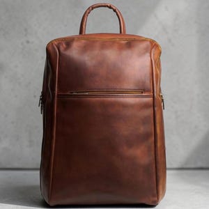 Puede incluir: Una mochila de cuero marrón estructurada con un acabado mate liso, un asa superior única y un bolsillo horizontal con cremallera en la parte superior frontal. El diseño incluye costuras verticales limpias a lo largo de los lados y forma rectangular.