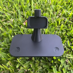 Può includere: Un espositore in plastica nero opaco caratterizzato da una base rettangolare con due rientranze circolari, un supporto centrale conico e una piccola piattaforma rettangolare superiore progettata per sostenere un oggetto in posizione verticale.
