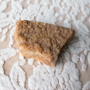 May include: A rough, irregularly shaped piece of brown and tan rock with a crystalline texture. The rock is sitting on a white lace surface.