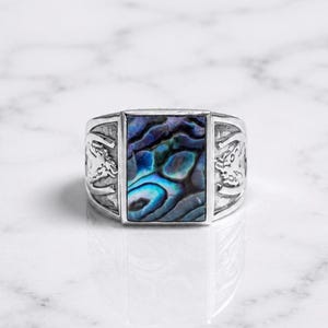 May include: A silver ring featuring a rectangular abalone shell inlay with iridescent blue, green, and purple tones. The band has a polished silver finish with detailed embossed horse motifs on both sides of the central shell setting.