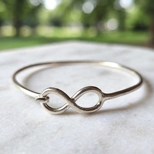 Può includere: Un bracciale rigido in metallo color argento caratterizzato da un simbolo dell'infinito centrale. Il bracciale presenta una finitura liscia e lucida e un cinturino circolare continuo che si collega all'anello dell'infinito sulla parte anteriore.