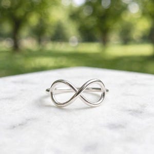 May include: A polished silver-toned ring featuring a prominent infinity symbol design centered on the band. The metal surface is smooth and reflective, showcasing a continuous loop shape that forms the focal point of this simple jewelry piece.