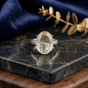 May include: A silver ring featuring a large, oval-cut rutilated quartz gemstone with visible golden needle-like inclusions. The band has a textured, ribbed design on the sides, providing a distinct contrast to the smooth, faceted surface of the stone.