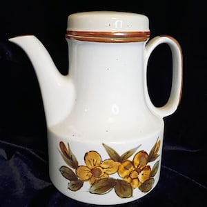 May include: A ceramic coffee pot with a white speckled glaze, featuring a brown-rimmed lid, a curved handle, and a spout. The base is decorated with a hand-painted floral motif in shades of yellow, brown, and green against the white body.
