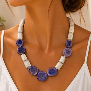 Puede incluir: Un collar llamativo que presenta una alternancia de cuentas cilíndricas blancas y rodajas circulares de piedra druzy morada. El diseño incorpora pequeños espaciadores plateados entre las cuentas, creando un patrón estructurado.