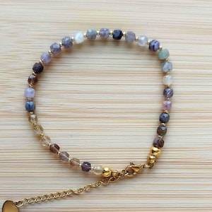 Può includere: Un braccialetto delicato con piccole perle di pietre preziose sfaccettate multicolori nei toni del viola, blu, marrone e trasparente, intervallate da minuscole perline distanziatrici in metallo dorato. Chiusura a moschettone in metallo dorato.