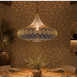 May include: A metallic pendant light fixture with a wide, bell-shaped shade featuring intricate, laser-cut geometric patterns. The gold-toned metal surface allows light to pass through the cutouts, casting patterned shadows on the surrounding walls.
