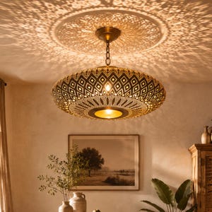Pode incluir: Uma luminária pendente redonda de metal com acabamento em bronze escuro e padrões geométricos intrincados cortados a laser. A luminária é suspensa por uma corrente curta e projeta uma luz complexa em forma de sombra no teto.
