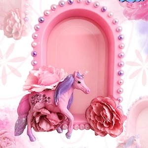 Könnte beinhalten: Ein rosafarbener, bogenförmiger Präsentationsrahmen, verziert mit einer Bordüre aus kleinen irisierenden Perlen. Eine rosa Einhornfigur mit violetter Mähne und Schweif steht davor, geschmückt mit großen rosa Stoffrosen und dem Wort MAGIC in Buchstaben.