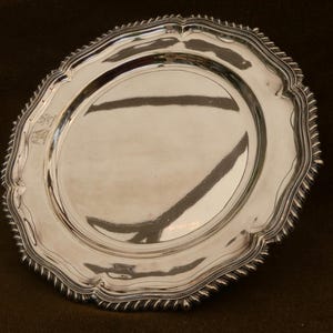 May include: A polished silver-toned serving platter with a smooth, reflective circular centre and a decorative, scalloped edge featuring a rope-like texture. The metal surface displays subtle reflections and a small engraved hallmark on the rim.