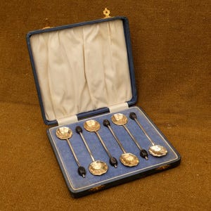 May include: A set of five silver-toned spoons with scalloped bowls and black oval accents on the handles, presented in a blue hinged case with a cream-coloured satin interior lining. The spoons are arranged in a row within the blue velvet base.