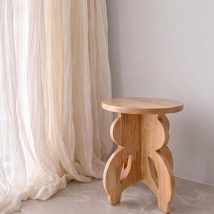 May include: A light-toned wooden stool featuring a circular top and a base constructed from three interlocking vertical panels with curved, cutout edges. The stool has a smooth, natural wood grain finish and stands on a flat, neutral-colored surface.