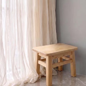 May include: A small, rectangular wooden stool with a light natural finish. The stool features four sturdy legs connected by horizontal support bars on all sides, providing a stable base. The top surface is smooth with rounded corners and edges.