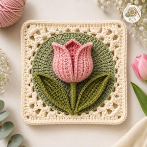 Puede incluir: Un motivo de ganchillo cuadrado que presenta un círculo central verde oliva con una flor de tulipán rosa tridimensional y dos hojas verdes. El cuadrado está enmarcado por un borde de encaje color crema con un patrón geométrico repetitivo.