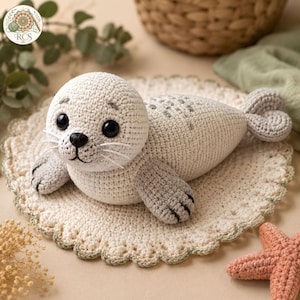 Puede incluir: Un juguete de peluche de foca hecho a ganchillo en color crema con ojos oscuros, nariz negra y aletas con puntas grises. La foca presenta manchas grises bordadas en la espalda y bigotes blancos, descansando sobre un tapete de ganchillo redondo.
