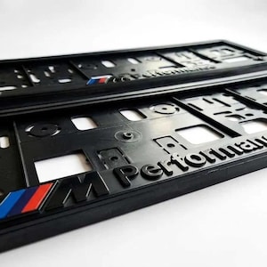Può includere: Due portatarga in plastica nera con il testo in rilievo M Performance e l'iconica striscia con logo M blu, viola e rossa sul bordo inferiore. Questi telai rettangolari presentano molteplici fori e fessure di montaggio per l'installazione.