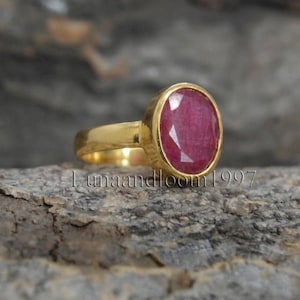 May include: A gold ring featuring a large, oval-cut, deep pink gemstone set in a smooth metal bezel. The band is polished gold, and the stone displays a faceted surface that reflects light. The text "lunaandloom1997" is visible across the image.