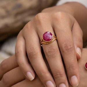 Könnte beinhalten: Ein goldener Ring mit einem runden, facettierten, tief rosa-roten Edelstein in einer Zargenfassung. Die Ringschiene ist in zwei dünne, parallele Golddrähte geteilt, die an der Basis der Fassung zusammenlaufen und ein schlichtes Design bilden.