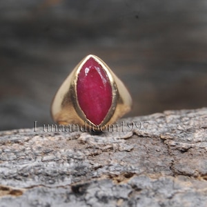 Peut inclure: Une bague en or avec une pierre précieuse rubis rouge foncé taille marquise dans un serti clos lisse. Le texte "lunaandloom1997" est visible au centre de l'image, superposé sur la bague et la surface texturée située derrière celle-ci.
