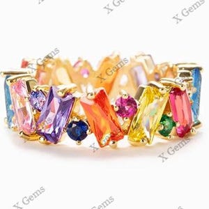 Puede incluir: Un anillo de tono dorado con una serie de piedras preciosas de colores en talla baguette y redonda. Las piedras están dispuestas en una banda continua, mostrando tonos vibrantes de morado, naranja, amarillo, rosa, azul y verde en un marco.